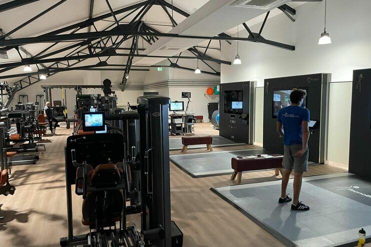 Fitnessraum mit mehreren Trainingsgeräten, einem jungen Mann in blauer Kleidung und Monitoren an den Wänden.