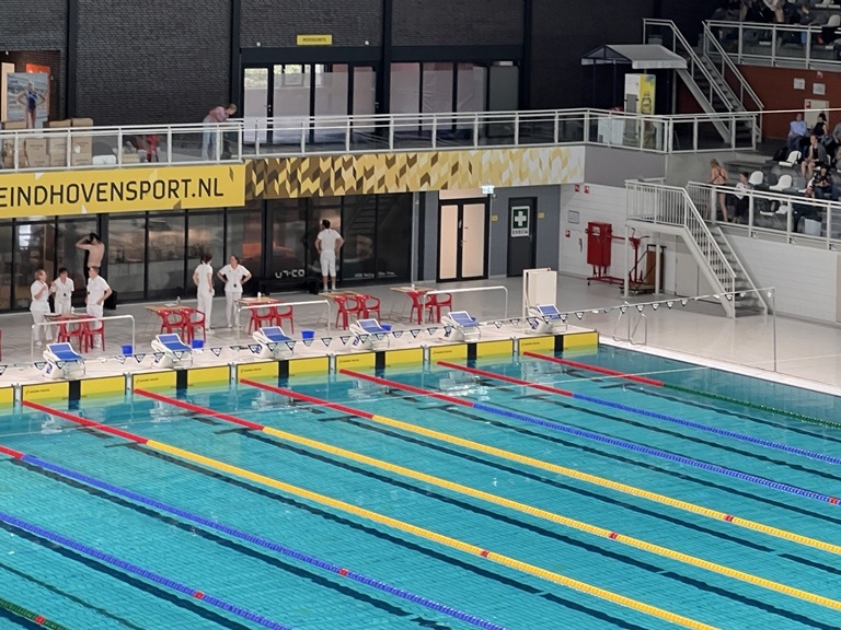 Schwimmhalle mit blauem Wasser, Startblöcken und Besuchern; Anzeige "Pieter van den Hoogenband" im Hintergrund.