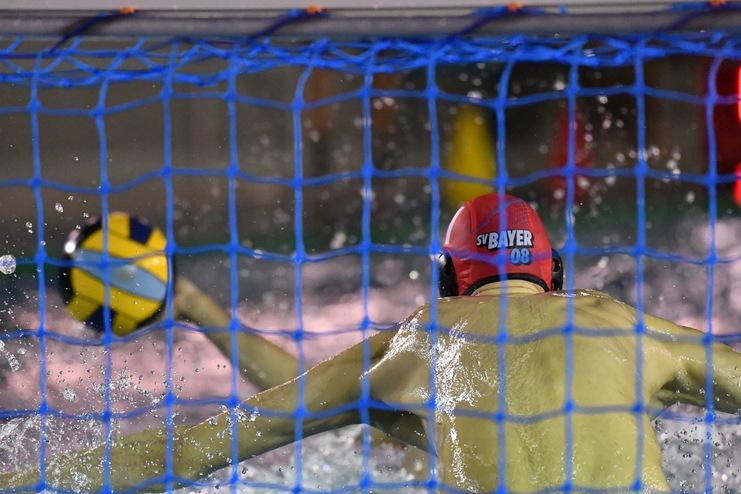Wasserballspieler wirft einen Ball durch das blaue Netz eines Torraums, umgeben von spritzendem Wasser.