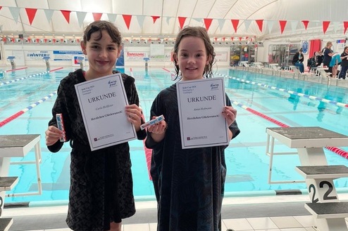 Zwei junge Schwimmerinnen in Bademänteln halten Urkunden neben einem Schwimmbecken in einer Sporthalle.