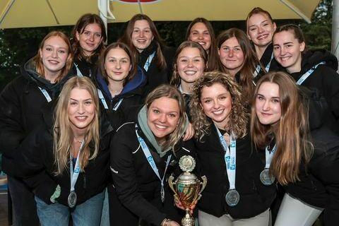 Gruppenfoto von zwölf Mädchen mit Medaillen, die einen Pokal in der Mitte stolz halten, vor einem überdachten Außenbereich.