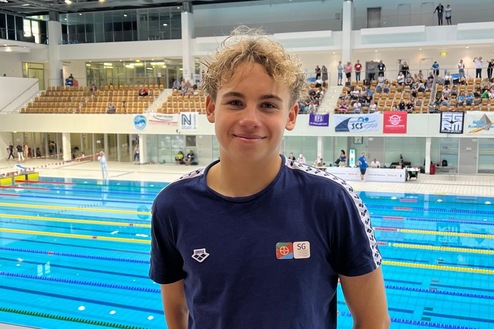 Junger Schwimmer steht lächelnd vor einem Wettbewerbspool, Umkleide im Hintergrund, Tribünen mit Zuschauern.