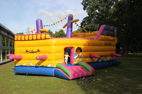 Buntes Hüpfburgen-Schiff mit Eingang, umgeben von Rasen, unter blauem Himmel und bunten Fähnchen.