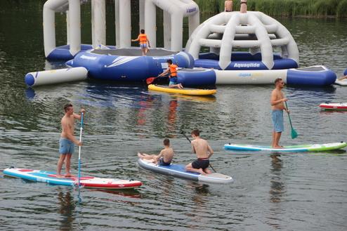 Mehrere Personen auf Stand-Up-Paddle-Boards und einem Wasser-Spielplatz in einem See, sonniger Tag.