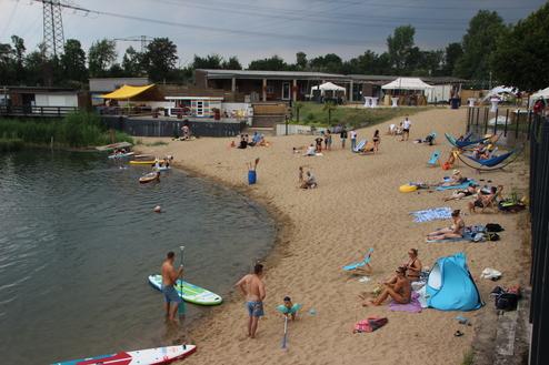 Strandaufenthalt mit Badegästen, Stand-Up-Paddle-Boardern und Sonnenschirmen an einem See. Sandstrand und grüne Umgebung.
