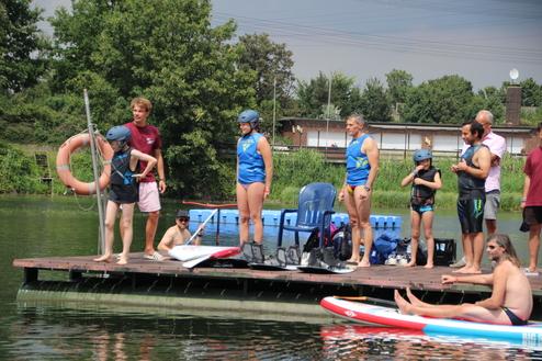 Gruppenszene auf einem Badeplatz: Personen in Schwimmbekleidung stehen auf einem Holzsteg am Wasser, einer paddelt im Hintergrund.