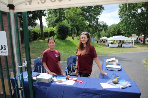 Zwei Frauen in roten T-Shirts stehen an einem Tisch, umgeben von einer grünen Wiese und Veranstaltungszelten im Hintergrund.