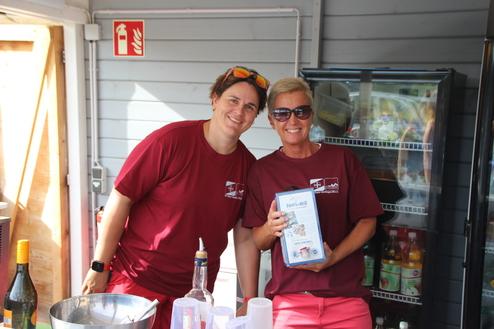 Zwei Frauen in roter Freizeitkleidung stehen lächelnd hinter einem Thekenbereich und halten eine Verpackung in der Hand.
