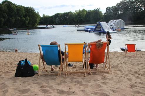 Drei Liegestühle am Sandstrand, Blick auf einen See mit Wasserattraktionen und Personen beim Stand-up-Paddeln.