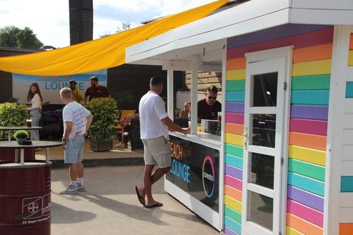 Bunte Beach Lounge mit Personen am Tresen, Pflanzenelementen und einer gelben Sonnenschutzplane im Hintergrund.