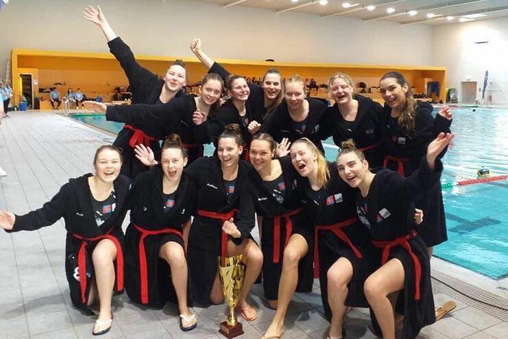 Gruppe von Jugendlichen in schwarzen Bademänteln mit Pokal, lächelnd am Beckenrand eines Schwimmbads.