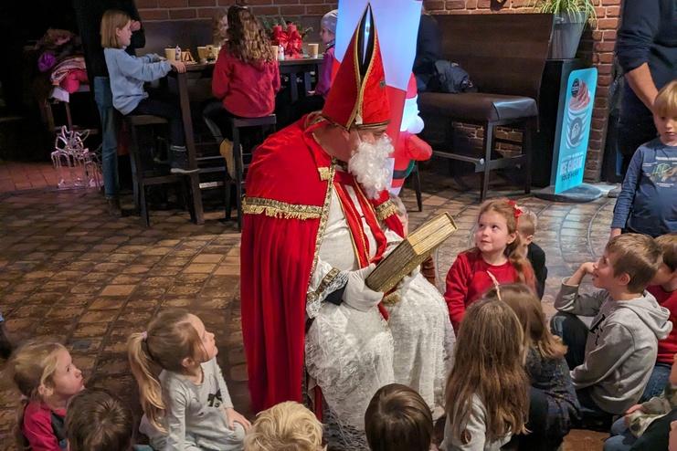 Der Nikolaus sitzt auf einem Stuhl und liest aus einem Buch, umringt von aufmerksamen Kindern.