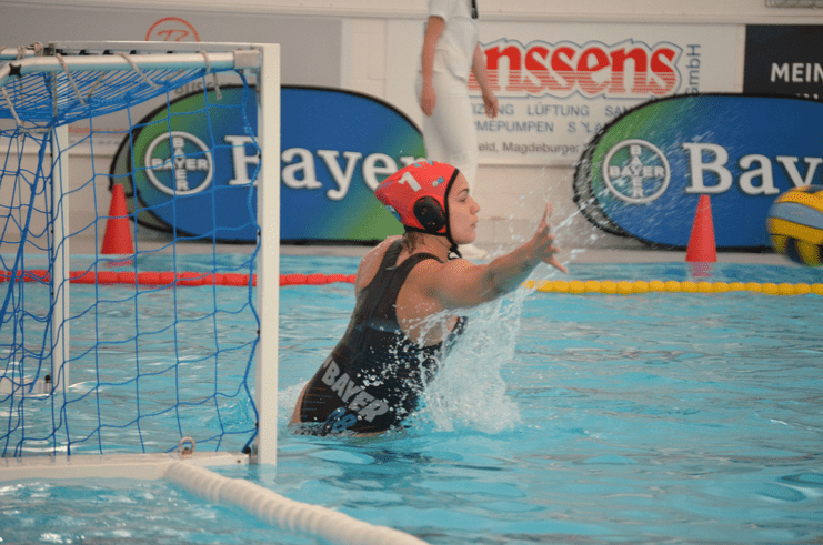 Wasserballspielerin wehrt einen Ball im Sprung vor dem Tor ab, Hintergrund mit Werbebannern und Wasseroberfläche.