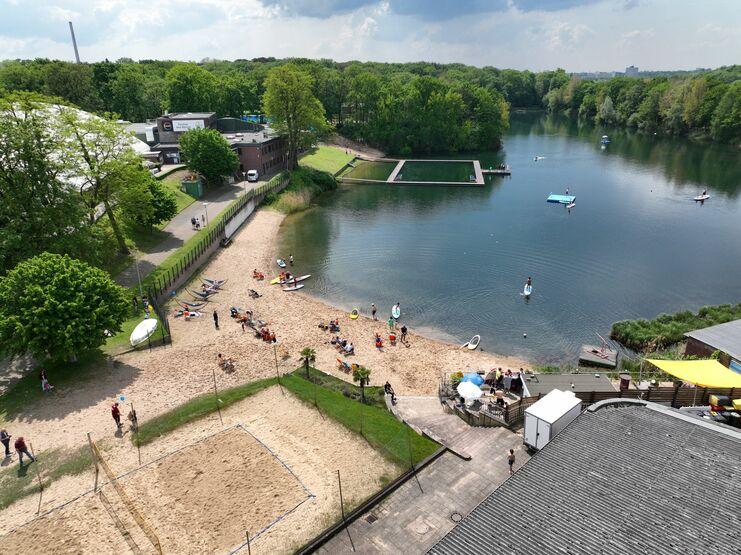 Luftaufnahme eines Gewässers mit Strand, Bootsverleih und Menschen beim Sonnenbaden und Wassersport.