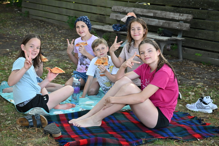 Fünf Kinder sitzen auf einer Decke im Freien und genießen Pizza, lächeln und zeigen Frieden-Zeichen.