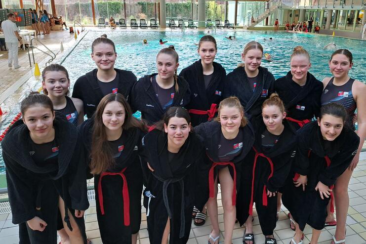 Gruppenumarmung von 14 jungen Schwimmerinnen in schwarzen Roben mit roten Gürtel am Beckenrand eines Schwimmbads.