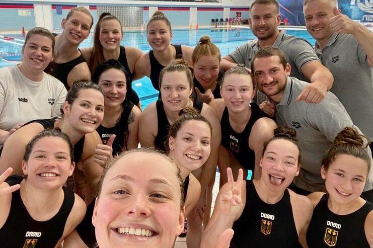 Gruppe von Wasserspringerinnen und Trainern l&auml;chelt in einer Sporthalle mit Schwimmbecken im Hintergrund.
