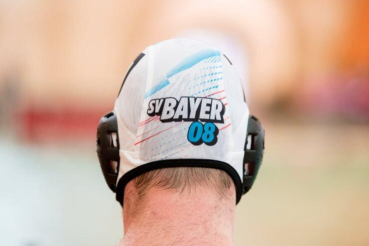 Nahaufnahme eines Schwimmers mit einer weißen Badekappe mit „SV Bayer 08“ und einem verschwommenen Schwimmbecken im Hintergrund.