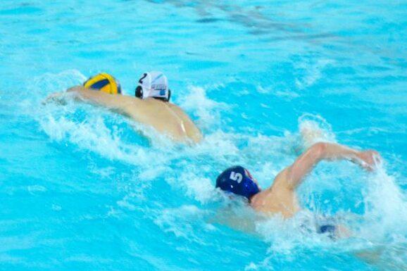 Zwei Wasserballspieler schwimmen in einem blauen Schwimmbecken, einer mit gelbem Ball, der andere mit blauer Kappe.