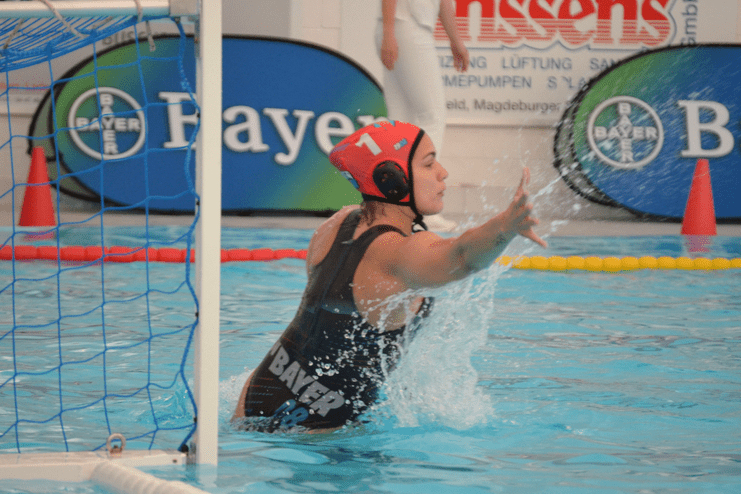 Wasserballspielerin wehrt einen Ball im Sprung vor dem Tor ab, Hintergrund mit Werbebannern und Wasseroberfl&auml;che.