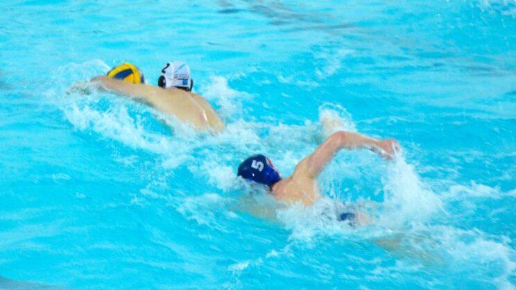 Zwei Wasserballspieler schwimmen in einem blauen Schwimmbecken, einer mit gelbem Ball, der andere mit blauer Kappe.