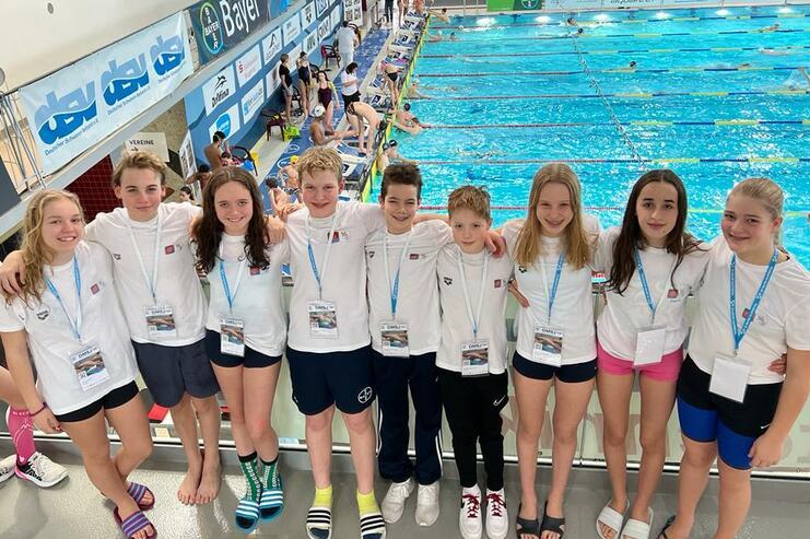 Gruppe von jungen Schwimmern in Weiß mit Medaillen am Hals, lächeln am Rand eines Schwimmbeckens.