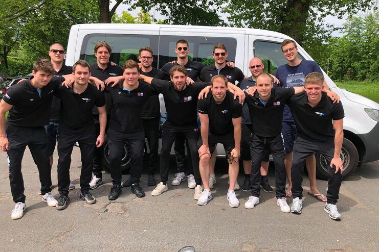 Gruppe von 15 Männern in schwarzen T-Shirts posiert vor einem weißen Kleintransporter auf einem Parkplatz.
