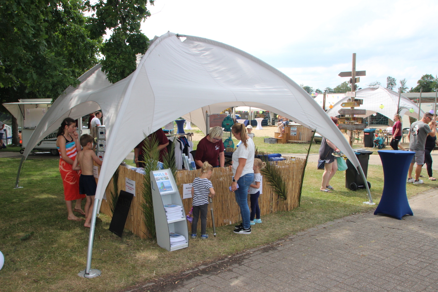 Wettbewerb mit Zelt, Leuten und Ständen auf einer Wiese, umgeben von grünen Bäumen und einem entspannten Veranstaltungsambiente.
