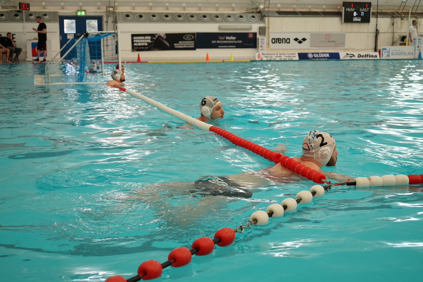 Zwei Wasserballspieler treiben im Becken, mit einem Tor und einer Abtrennungsleine im Hintergrund.