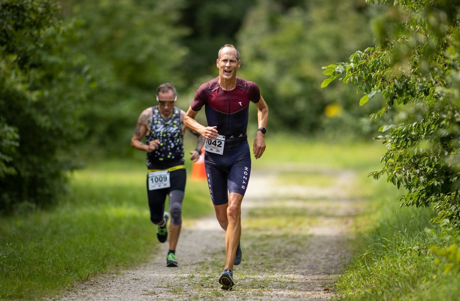 Zwei Athleten laufen auf einem Waldweg während eines Wettkampfs, einer trägt die Startnummer 1042, der andere 1009.