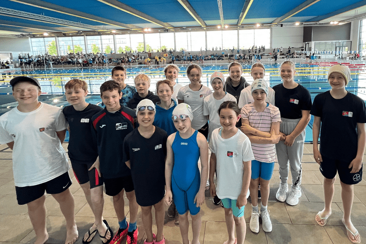 Gruppe von Schwimmern in Badebekleidung posiert am Beckenrand eines Schwimmzentrums mit blauen Decken und Zuschauern im Hintergrund.