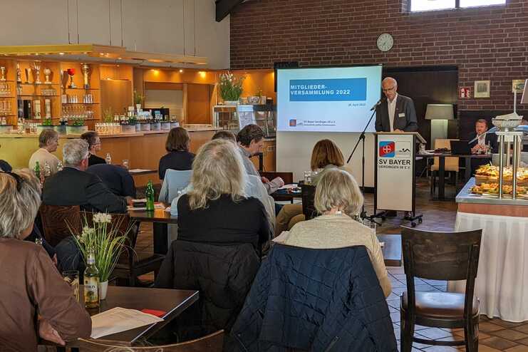 Mitgliederversammlung 2022 in einem Saal, mit Referent am Podium und Teilnehmern im Hintergrund. Buffet sichtbar.