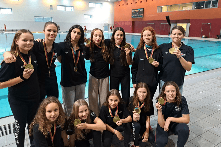 Gruppe junger Schwimmerinnen mit Goldmedaillen vor einem Schwimmbecken in einem Sportzentrum.