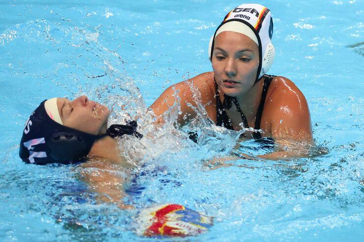 Zwei Wasserballspielerinnen k&auml;mpfen im Wasser, spritzendes Wasser um sie herum, ein Ball schwimmt nahe der Oberfl&auml;che.