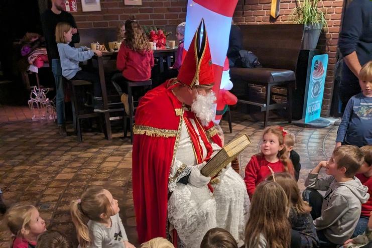 Der Nikolaus sitzt auf einem Stuhl und liest aus einem Buch, umringt von aufmerksamen Kindern.