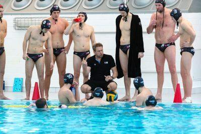 Wasserballmannschaft während einer Besprechung am Beckenrand; einige Spieler im Wasser, andere stehen um den Trainer.