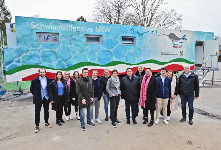 Gruppenfoto vor einem farbenfrohen Mobil der Schwimmoffensive NRW mit mehreren Personen in Freizeitkleidung.