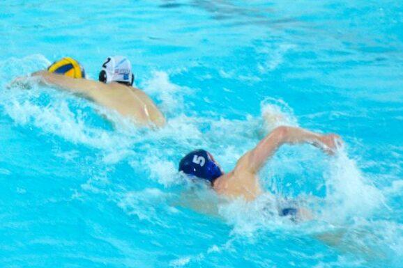 Zwei Wasserballspieler schwimmen in einem blauen Schwimmbecken, einer mit gelbem Ball, der andere mit blauer Kappe.