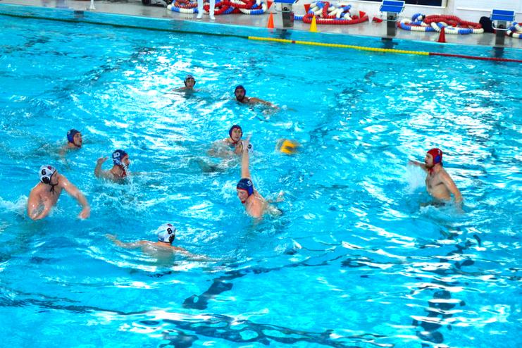 Wasserballspieler in Aktion in einem Schwimmbecken, w&auml;hrend einer Partie mit Ball und Tore im Hintergrund.