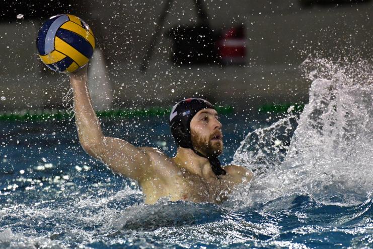 Ein Wasserballspieler mit einer gelb-blauen Kugel in der Hand spritzt Wasser beim Wurf in einem Schwimmbecken.