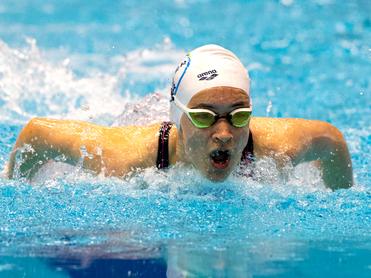 Schwimmerin in Spielhaltung mit Kappe und Brille, die intensiv im Wasser paddelt, spritzendes Wasser um sie herum.