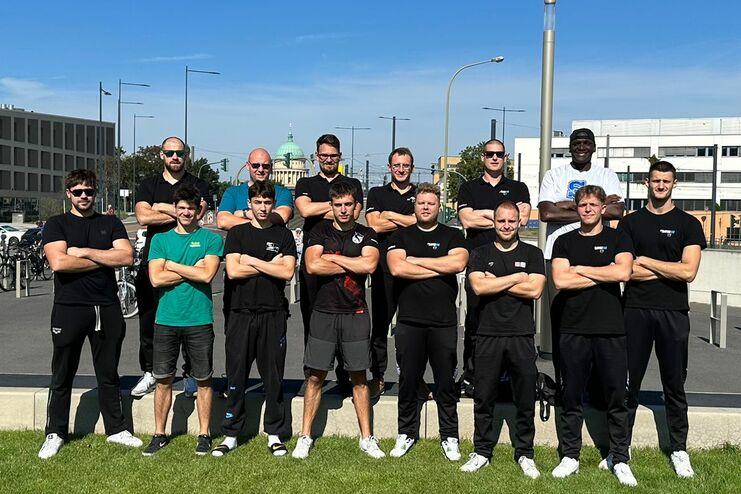 Gruppe von 15 Menschen in schwarzen T-Shirts posieren mit verschränkten Armen vor einem modernen Gebäude.