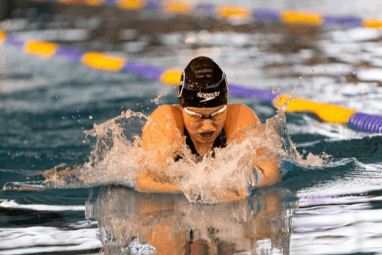 Schwimmerin in voller Aktion beim Brustschwimmen, mit Spritzwasser und bunten Bahnen im Hintergrund.