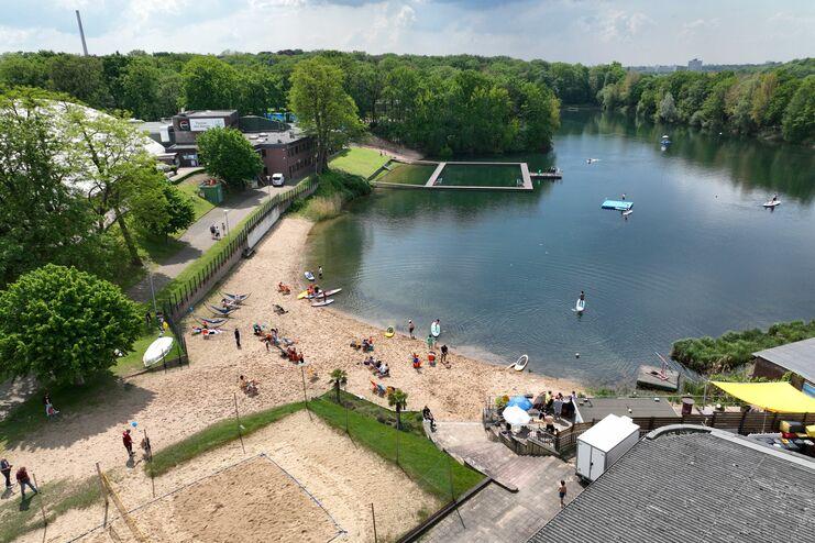 Luftaufnahme eines Gewässers mit Strand, Bootsverleih und Menschen beim Sonnenbaden und Wassersport.