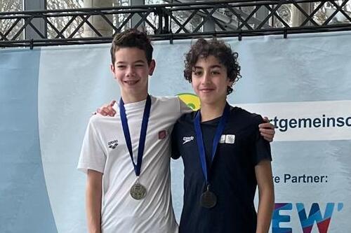 Zwei junge Sportler stehen auf dem Siegerpodest, tragen Medaillen und lächeln in die Kamera.
