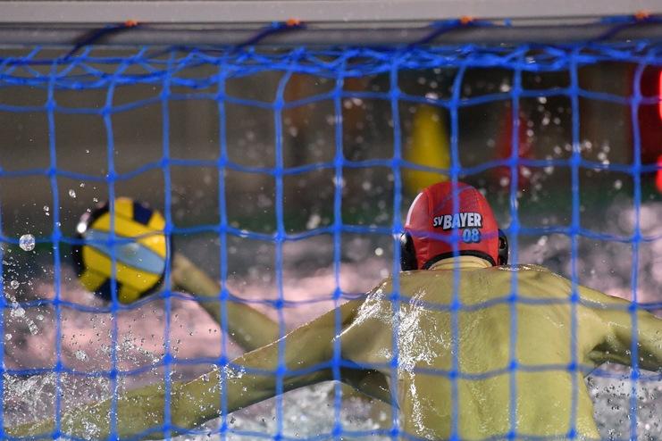 Wasserballspieler wirft einen Ball durch das blaue Netz eines Torraums, umgeben von spritzendem Wasser.