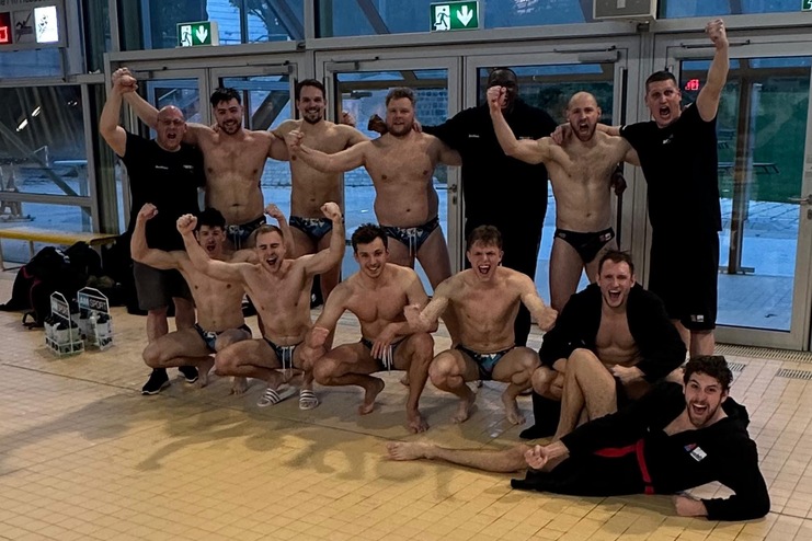 Gruppe von Männern in Badebekleidung posiert fröhlich in einem Schwimmbad mit großen Fenstern.