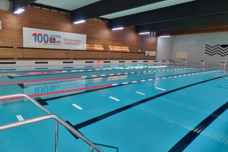 Innenansicht eines Schwimmbades mit einem Wettkampfbecken und einer Wandtafel, beschriftet mit „100 Jahre SV BAYER“.