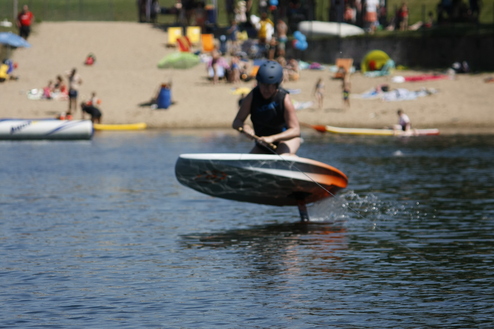 Junge f&auml;hrt auf einem Board &uuml;ber das Wasser, w&auml;hrend im Hintergrund ein Sandstrand mit Menschen sichtbar ist.