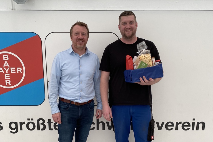 Zwei Männer stehen vor einer Wand mit dem Bayer-Logo, einer hält ein Geschenk, der andere lächelt.